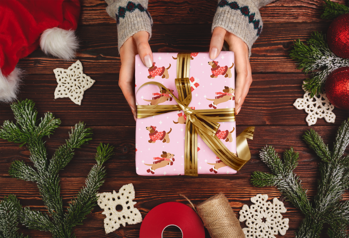 A top-down view of hands wrapped in gray knit sweaters holding a square gift box wrapped in pink paper with a gold ribbon and dog print. The scene is decorated with pine branches, white lace snowflakes, a red Santa hat, red ornaments, burlap, and red ribbon on a dark wooden table.